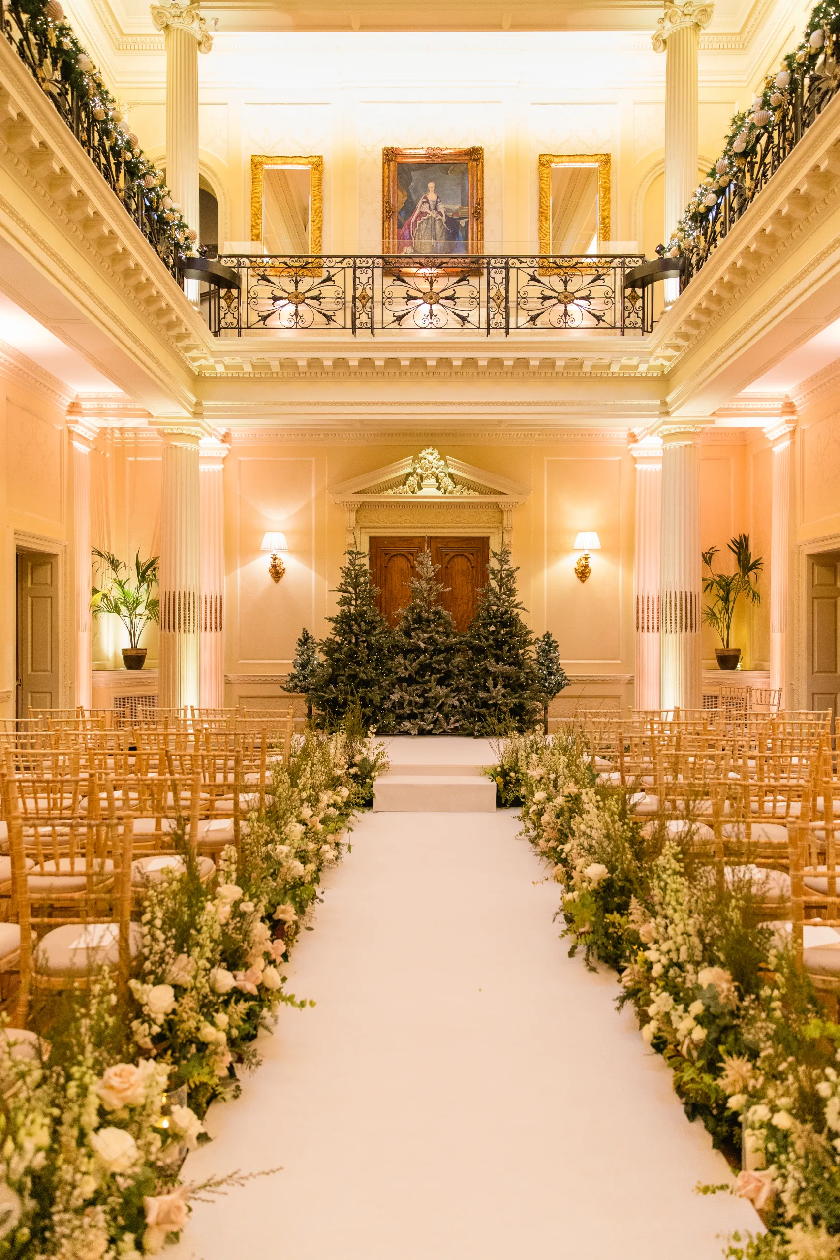 Interior of Hedsor House set up for wedding ceremony with floral displays at the end of the aisles and fir trees behind the stage.