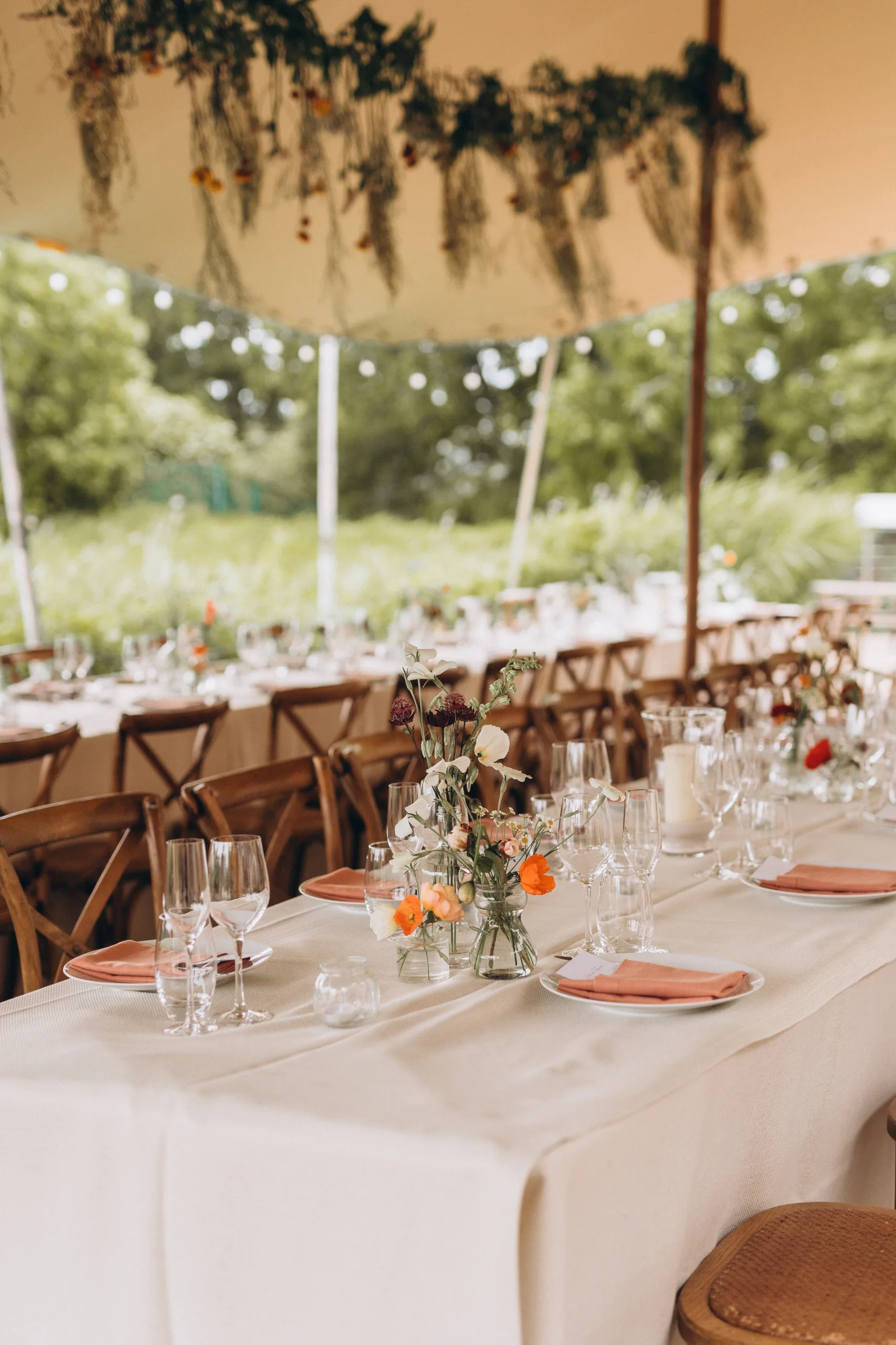 Wedding tables with white linen, place settings using salmon pink napkins and small jars of wildflowers, under an open marquee