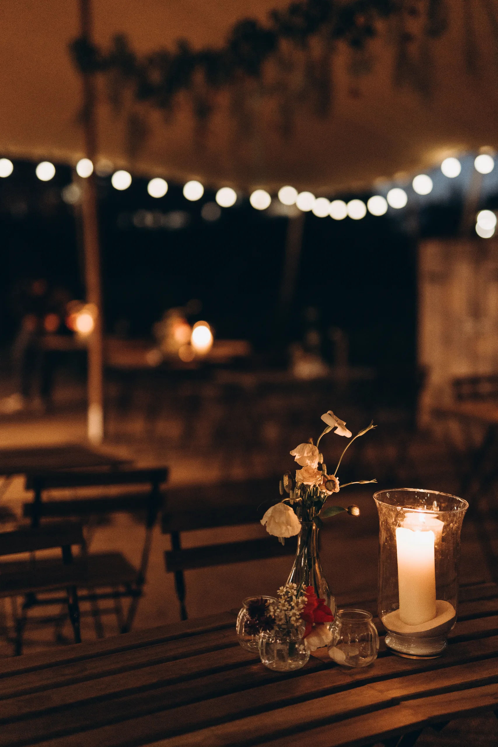 Pillar candle in glass jar next to vase with white flowers, on wooden table under marquee at night