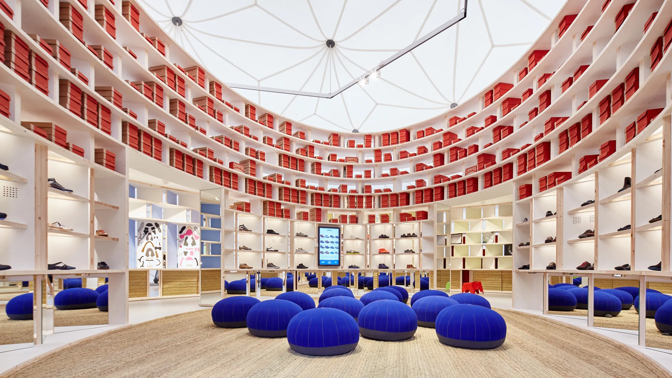 Pop-up shop at the Serpentine Gallery. The curved room is decorated in round shelves, some displaying shoes, others red shoe boxes, with round navy blue seats in the middle.
