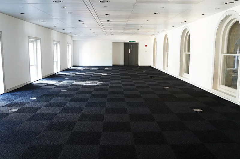 Empty gallery room at Old Billingsgate with chequered blue carpeting and white walls with windows on both sides
