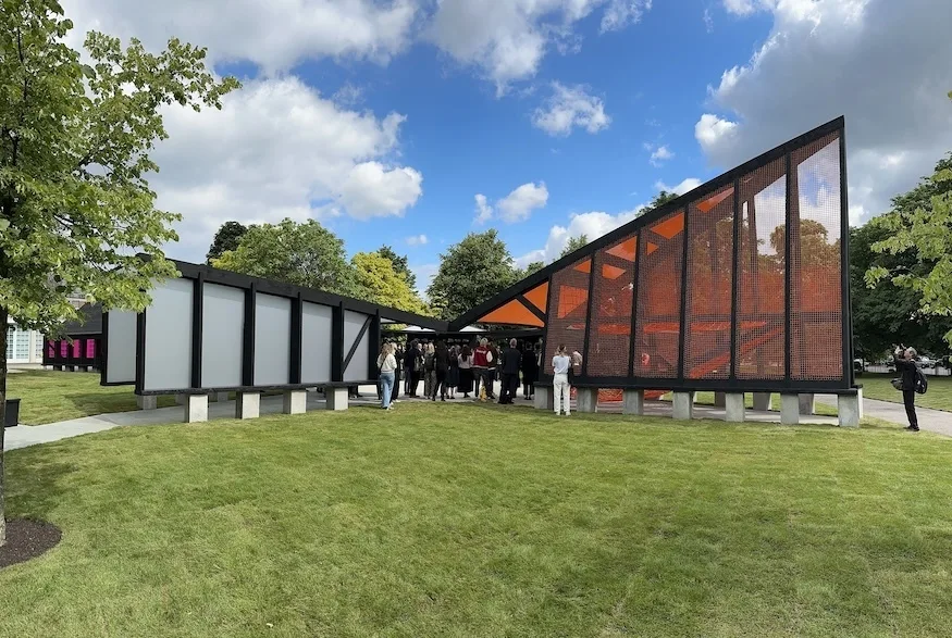 Pavillion at the Serpentine Gallery: a black frame structure with red glass triangle awning on one side and opaque white rectangular awning on the other