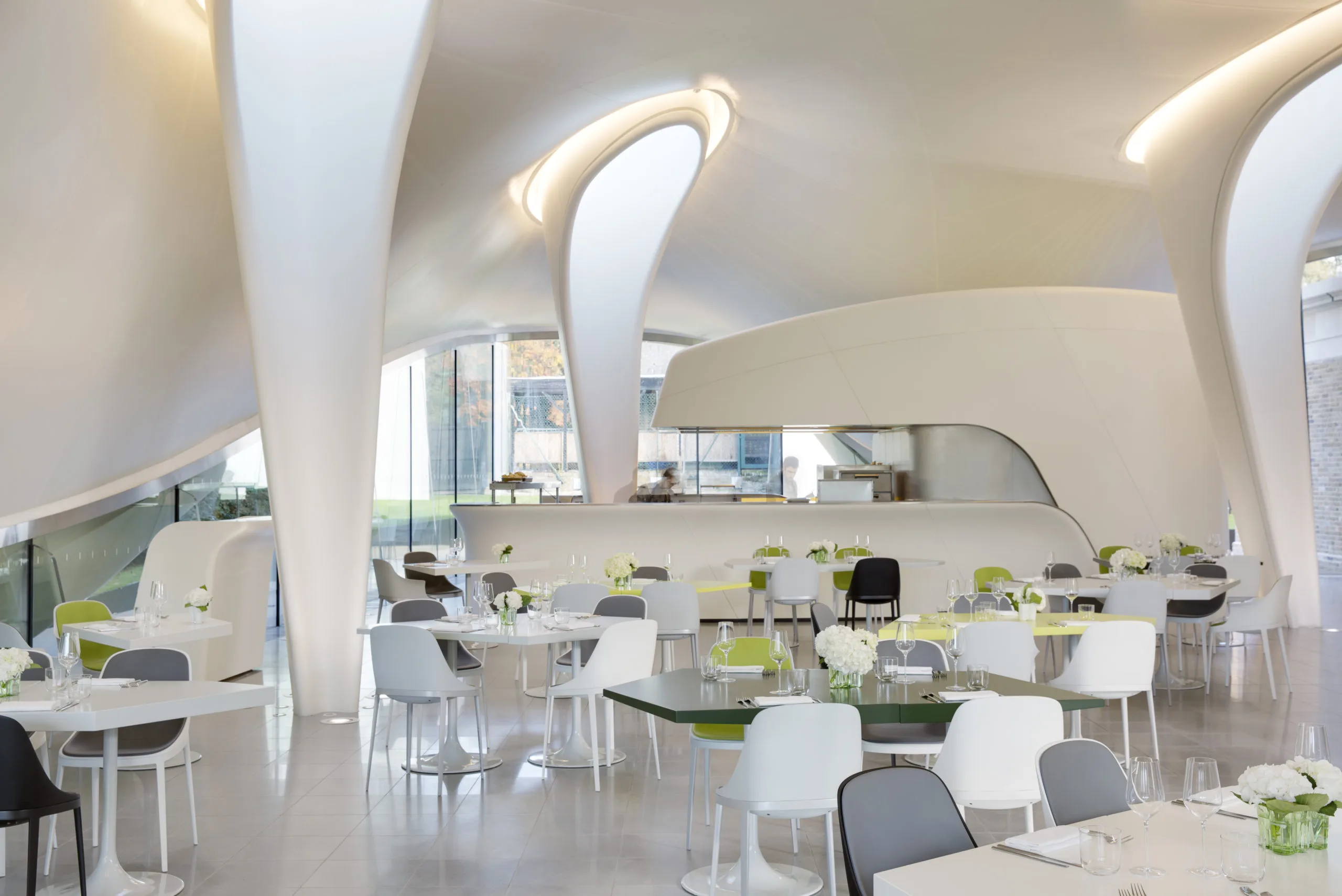 White dining room in sloping room with floor to ceiling white spoon-like shaped structures, glass dining tables and white chairs