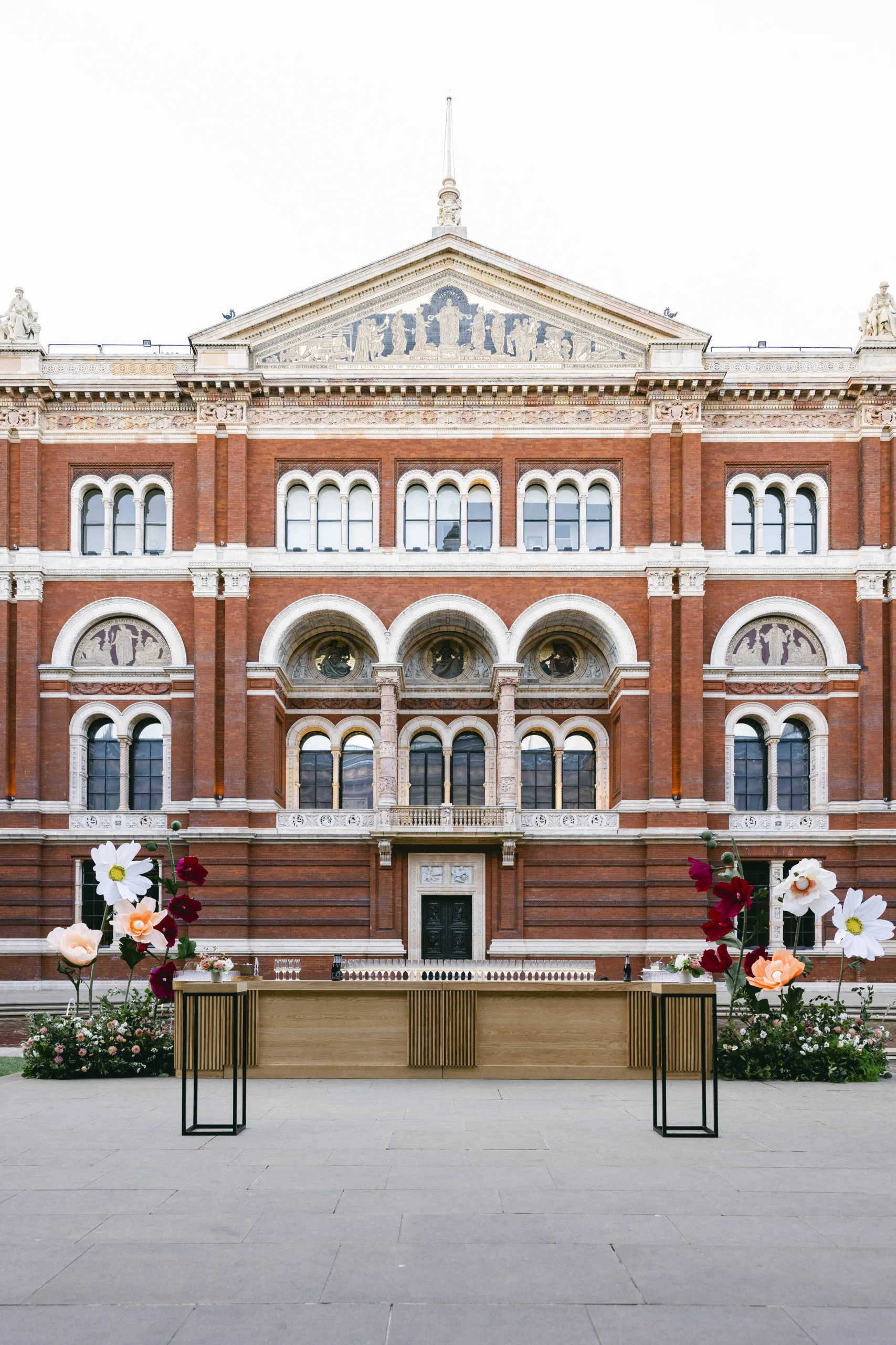 View of V&A facade, showing the full height of the building, with a display table of drinks set up in the courtyard alongside large flower installations.