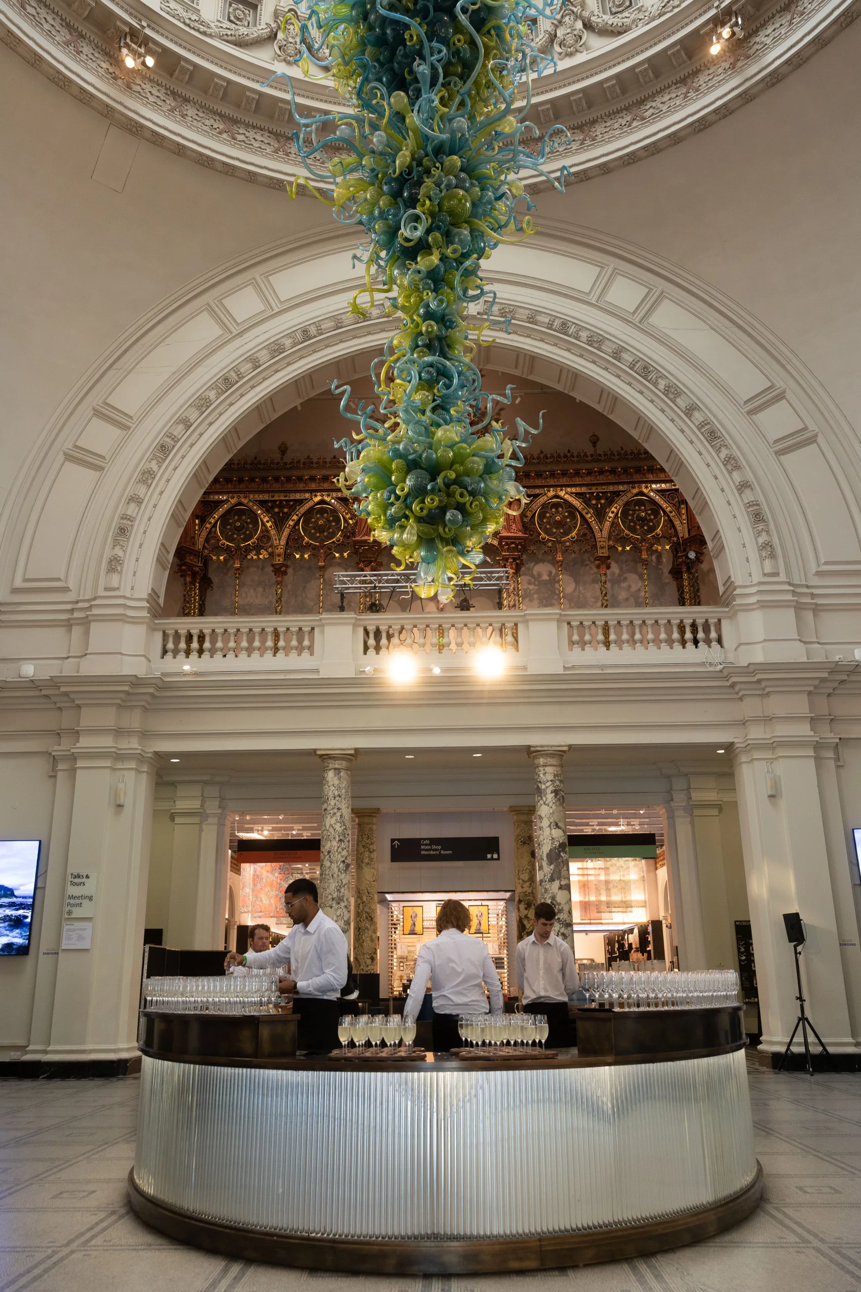 V&A reception area set up for drinks serve with waitstaff arranging glasses, under the large hanging glass structure.