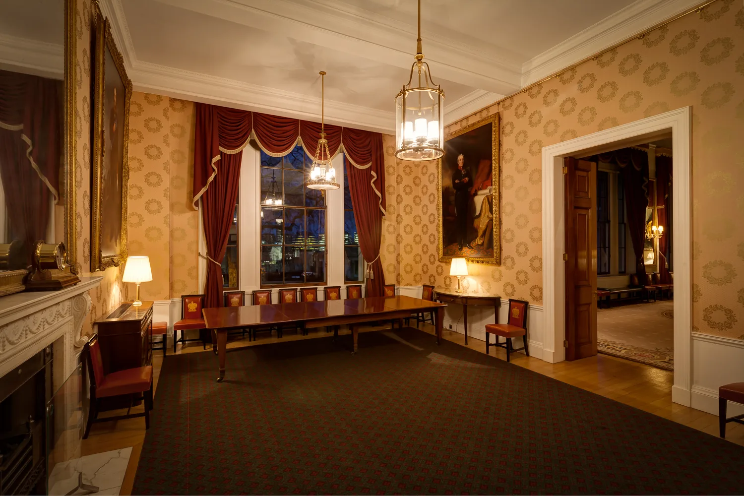 Yellow-walled room in Trinity House with wide wooden table at one side, with set of red chairs, red curtains hang around the window.