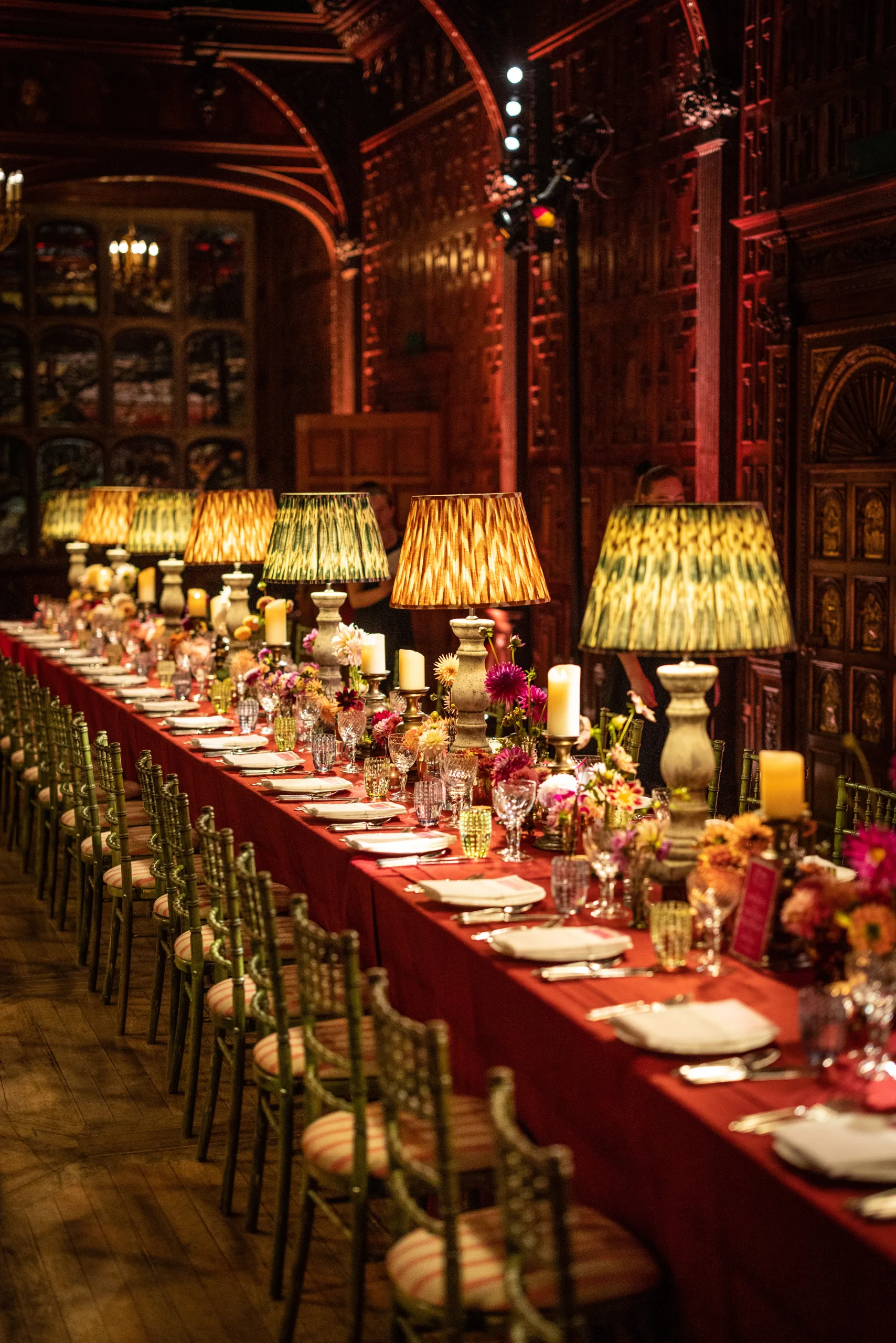 Long dining room table with rich red-lit room, set up for plate settings, glasses, candles on short candlesticks and vintage table lamps.